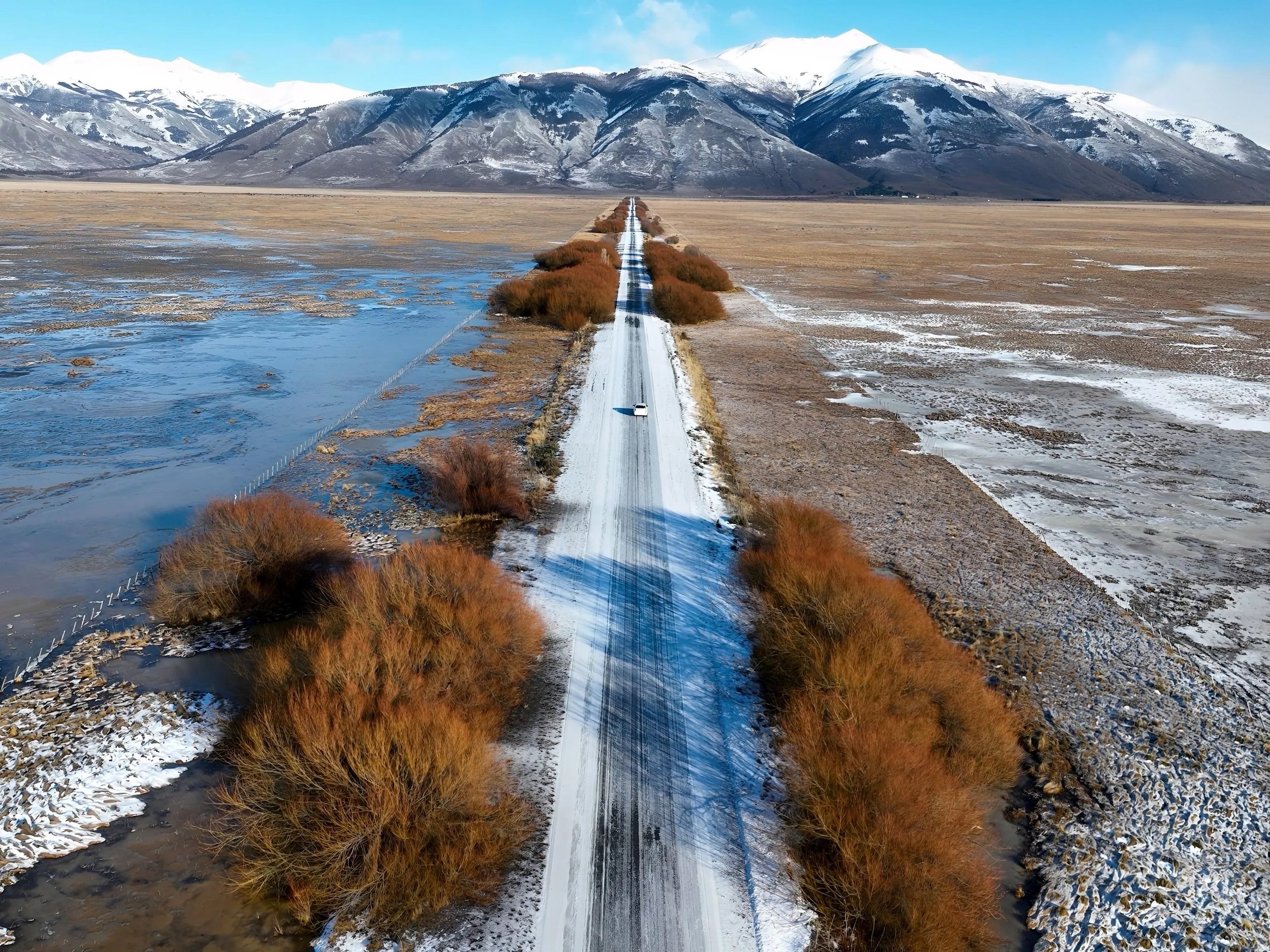 A long snow-covered road stretches toward snow-capped mountains in the distance, with patches of ice and sparse vegetation on either side.