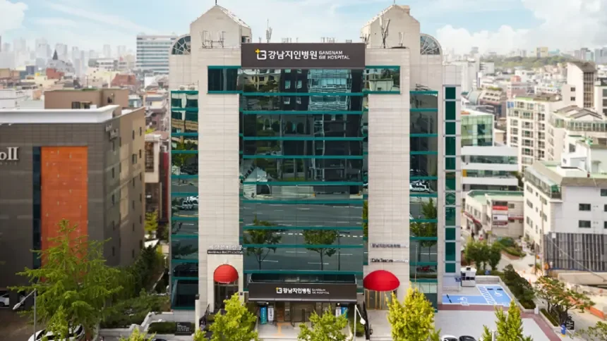 Modern multi-story building with reflective glass windows, sign reading GANGNAM GE HOSPITAL at the top, and red entrance awnings, surrounded by trees and other city buildings.