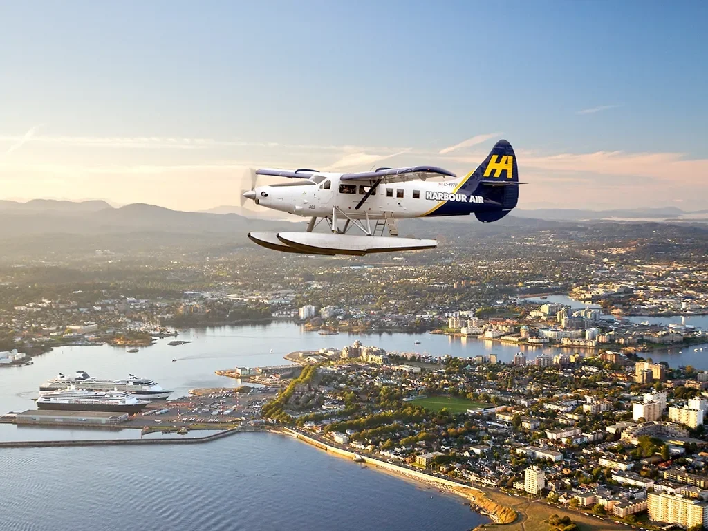 A seaplane flying over a city with a river and harbor, during sunset or sunrise.