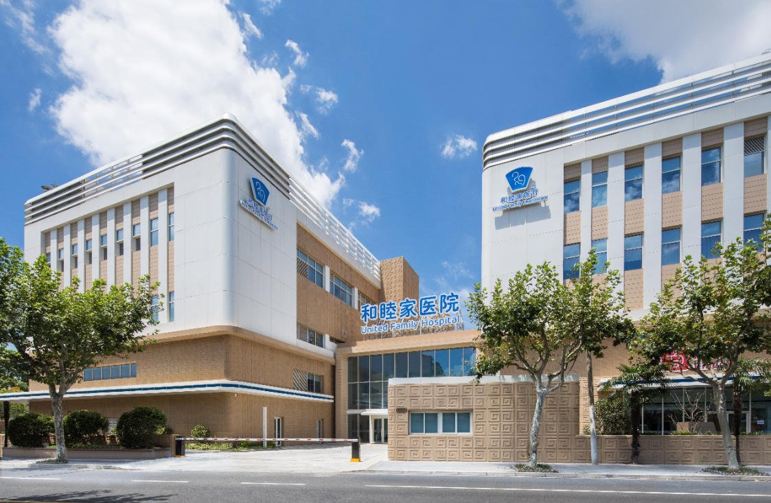 The front view of a large hospital building named United Family Hospital, with trees lining the sidewalk and a bright, partly cloudy sky overhead.