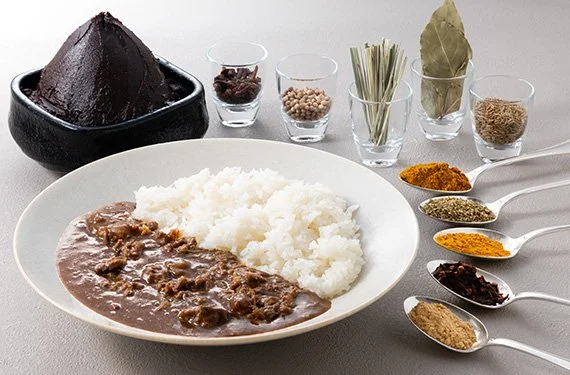 Plate of white rice and beef stew with spices in spoons, assorted spices in small bowls, and a black stone mortar filled with dark brown spice or paste.