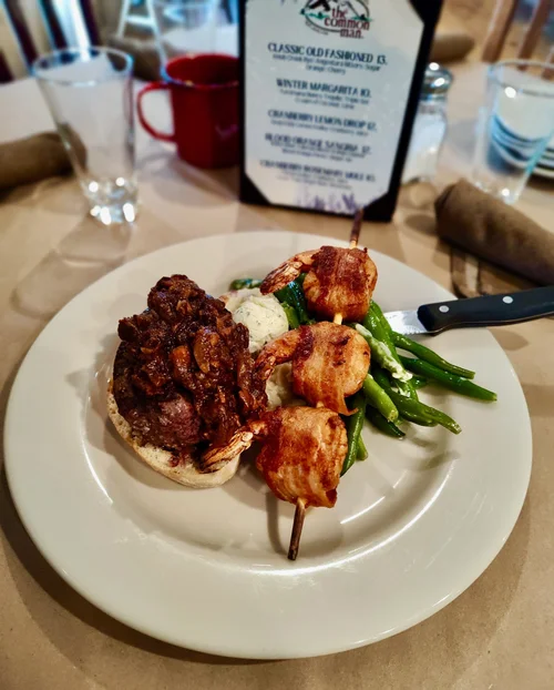 Plate with a meatloaf topped with a mushroom and onion mixture, green beans, and a skewer of bacon-wrapped shrimp, with a small scoop of butter or mashed potatoes.