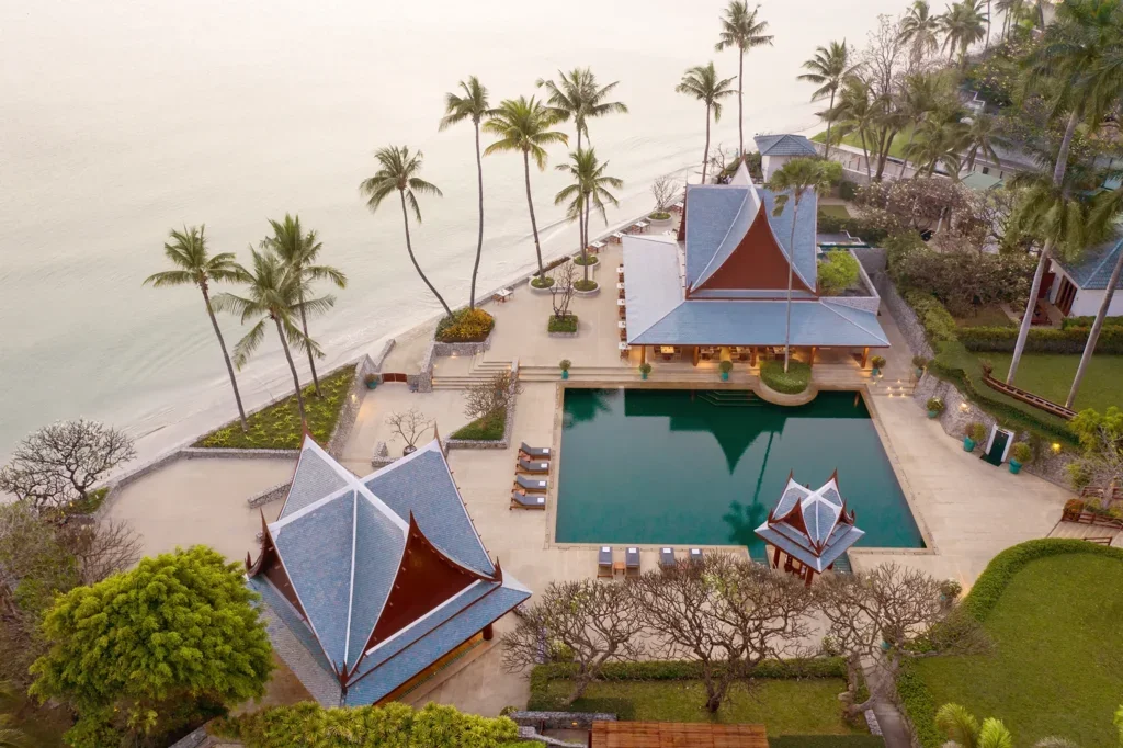 Aerial view of a tropical resort with a swimming pool, traditional Thai-style buildings, palm trees, and a beach shoreline.