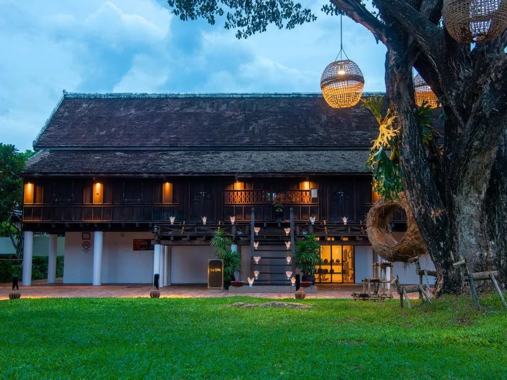 A traditional Thai wooden house on stilts with a staircase leading up to the main entrance, surrounded by a lush green lawn and large trees, with hanging lanterns and warm lighting at dusk.