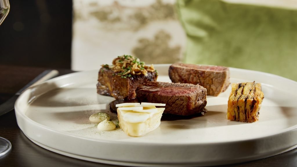 A plate with two pieces of cooked steak, a piece of layered vegetable terrine, a dollop of white sauce, and a small serving of mashed potatoes garnished with herbs.