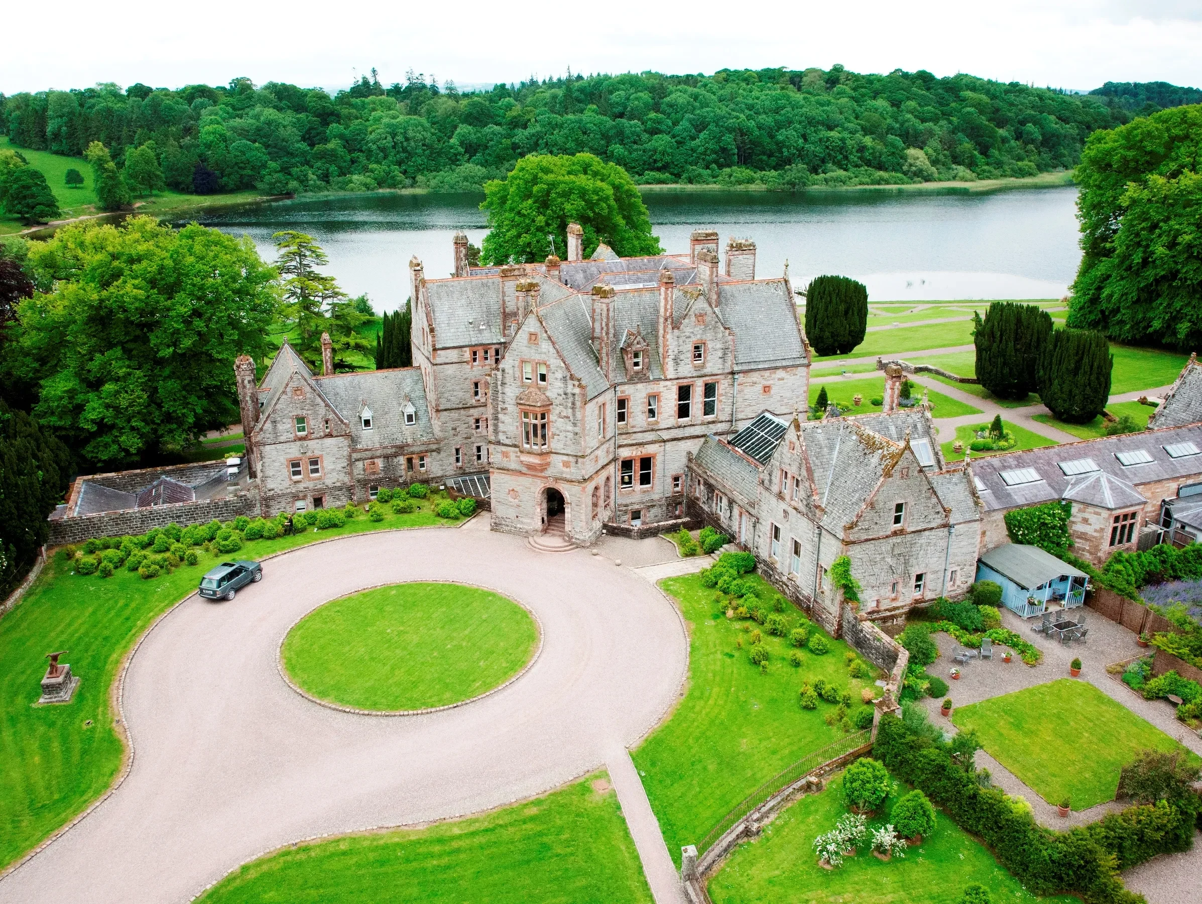 A large historic stone mansion surrounded by green trees, a well-maintained lawn, and a round gravel driveway in front, with a lake and forested hills in the background.