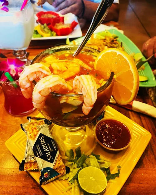 A seafood cocktail cocktail garnished with lemon slices and shrimp, served with a small cup of salsa, a packet of saltines, and a slice of lime, on a yellow plate. In the background, there are plates of food and drinks on a wooden table.