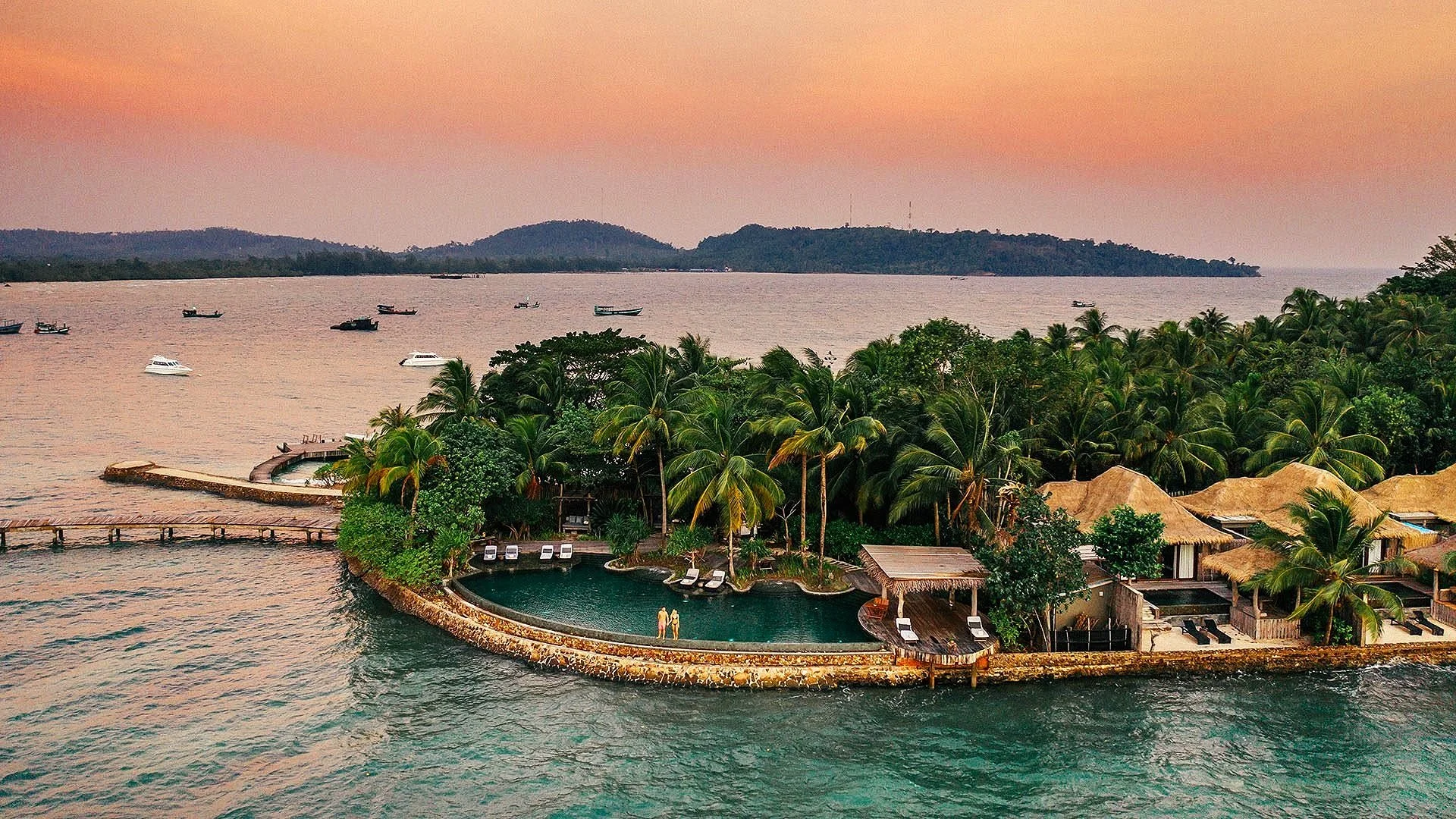 An aerial view of a tropical island with palm trees, bungalow-style buildings with thatched roofs, a swimming pool with two people, and boats on the water during sunset.