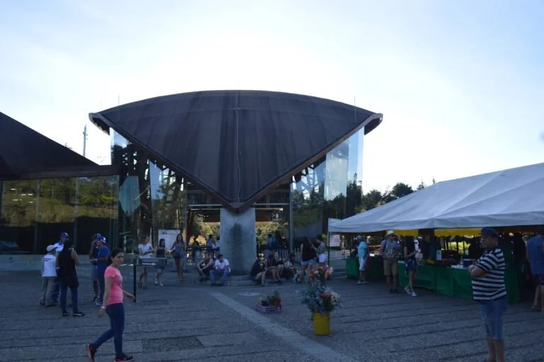 People gathered outside a modern building with a distinctive V-shaped roof and glass walls, some standing and sitting near flower arrangements and market stalls.