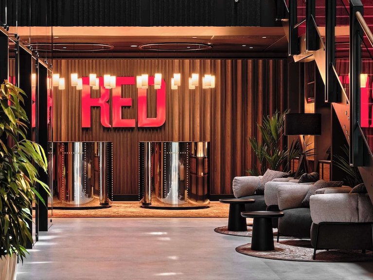Modern hotel lobby with a large illuminated "RED" sign on the back wall, wooden paneling, and comfortable seating area with gray sofas and black tables.