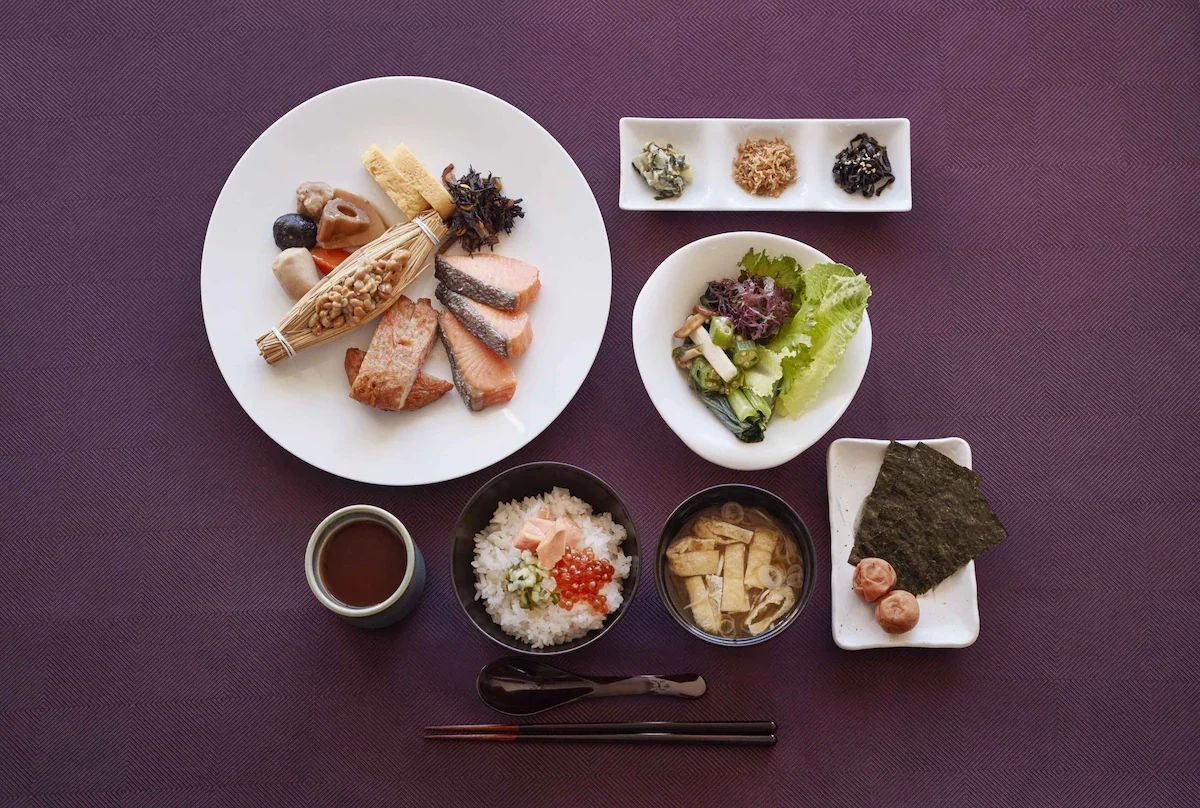 A traditional Japanese meal featuring grilled salmon, rice topped with salmon roe and pickled vegetables, miso soup with tofu and green onions, and various side dishes including seaweed snacks, pickled vegetables, and small appetizers on a purple tex