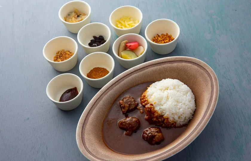 A plate of Japanese curry with white rice and several small bowls of assorted pickles, spices, and condiments on a blue table.