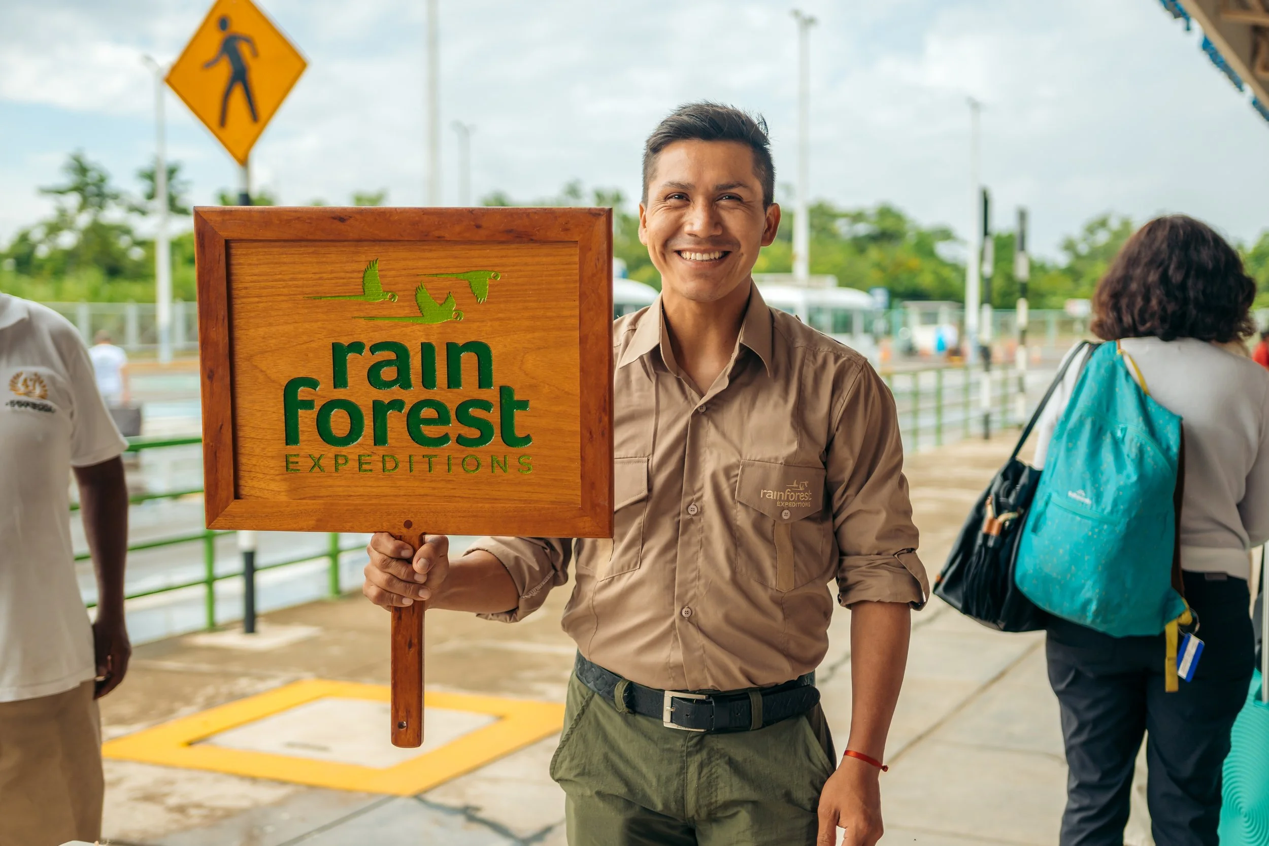 A smiling man in a beige shirt holding a wooden sign that reads "rain forest expeditions" with a green flying bird logo. He is standing outdoors near a green railing, with other people and trees in the background.