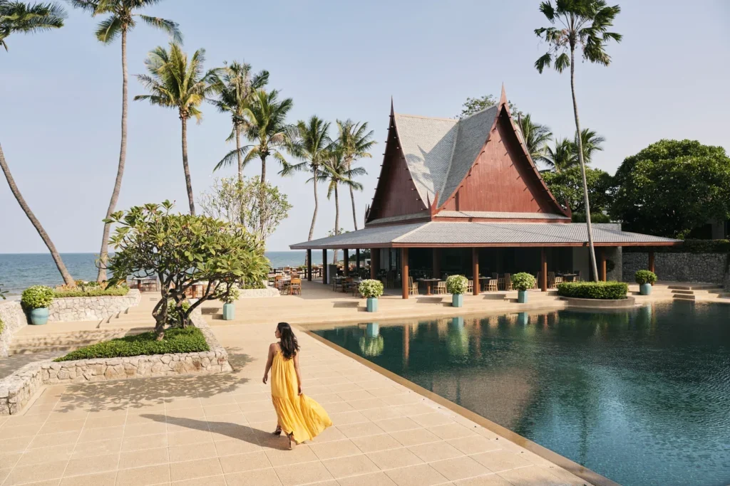 A woman in a yellow dress walking near a pool, with a traditional Thai-style building, palm trees, and the ocean in the background.