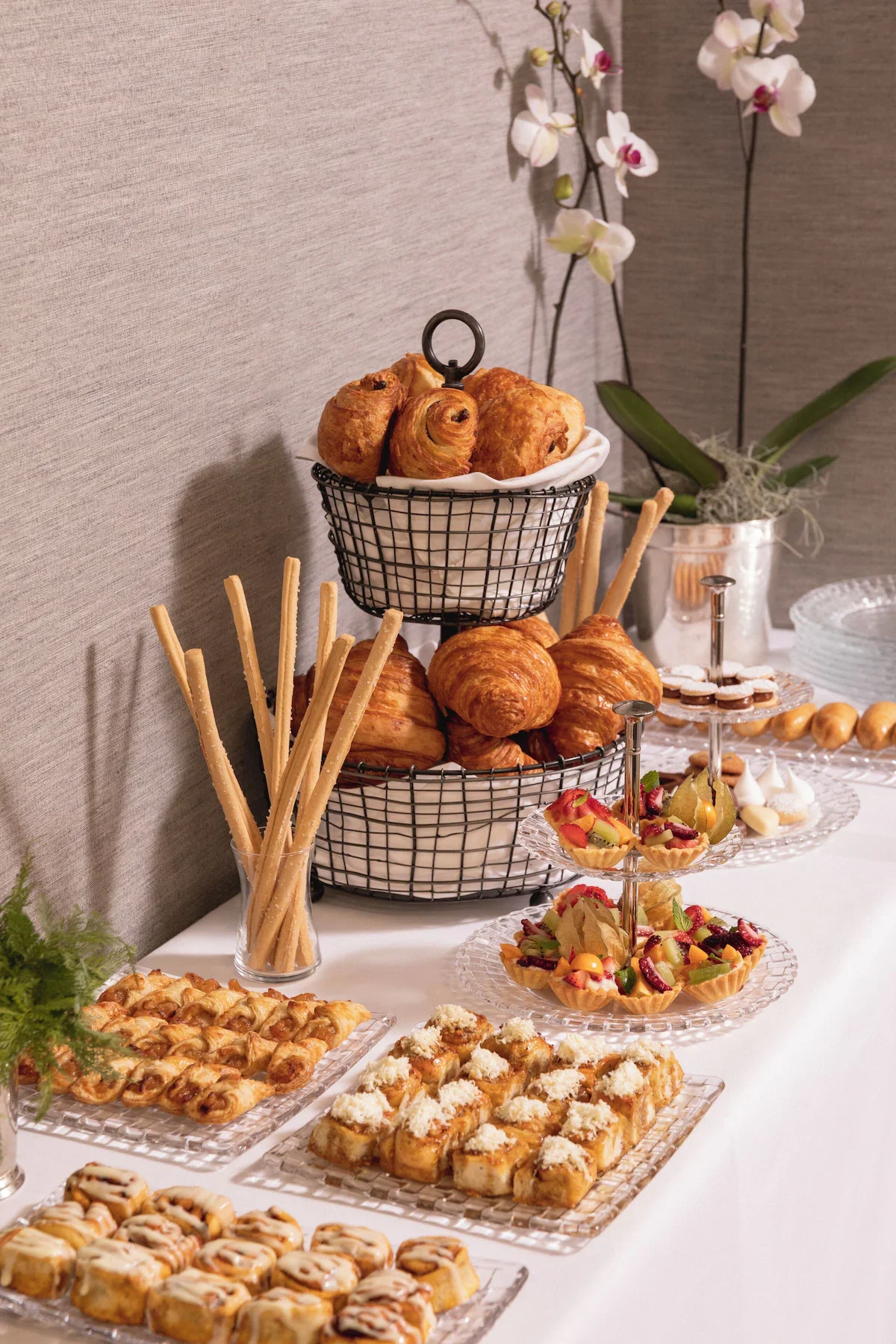 Assorted pastries, including croissants, danishes, and fruit tarts, arranged on a three-tiered tray and glass plates at a dessert table with a gray wall and an orchid in the background.