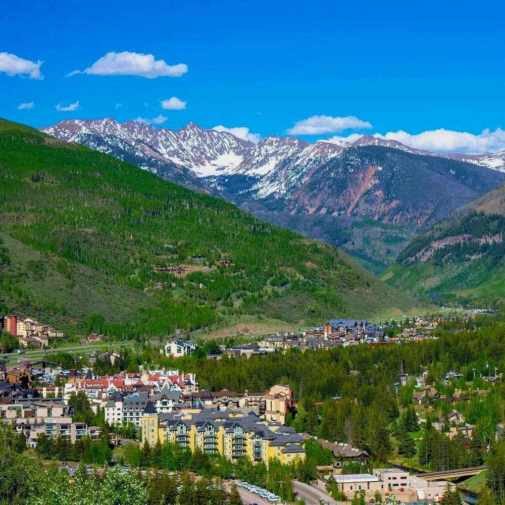 A mountain town with colorful buildings in a valley, green forested hills, and snow-capped mountains in the background under a blue sky with clouds.