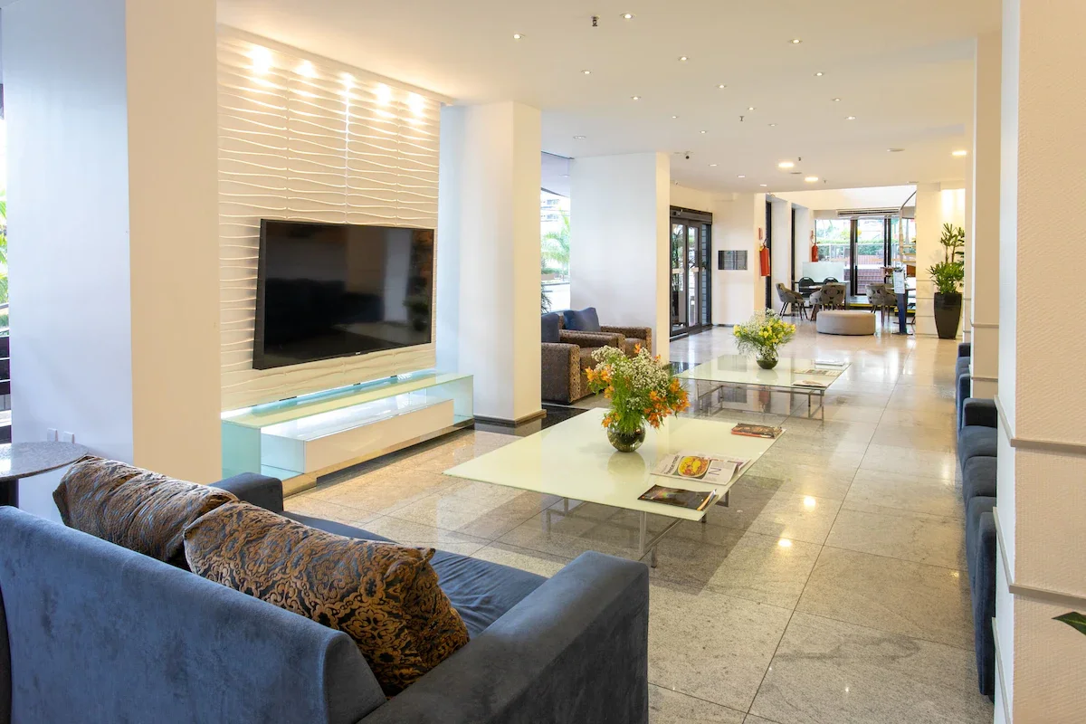 Modern hotel lobby with white walls, marble flooring, and a large flat-screen TV mounted on a textured wall. There are several seating areas with sofas and armchairs, glass coffee tables with floral arrangements and magazines, and large windows letti