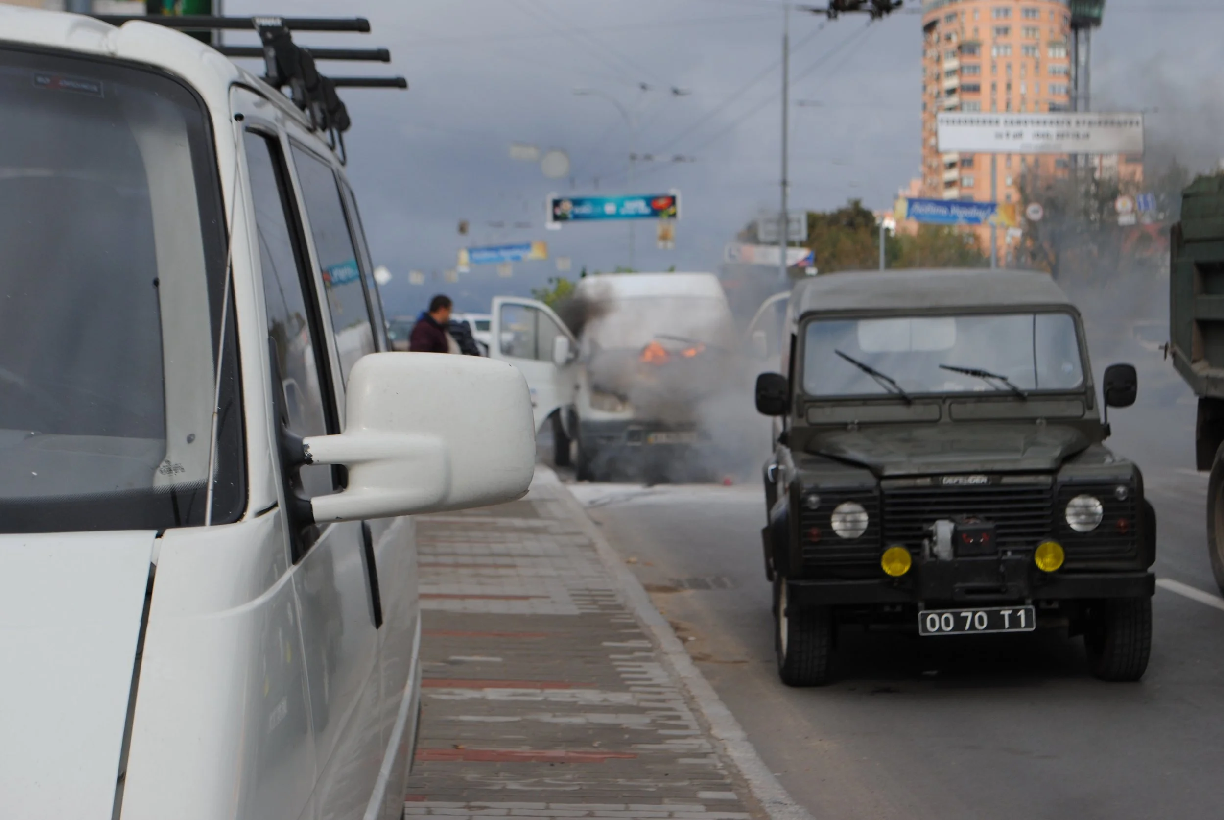 A traffic scene with several vehicles, including a white van and a small electric cart, on a city street under an overcast sky. Smoke or steam is rising from a burning vehicle in the background. Kiev November 2011, Marina Parrish ©