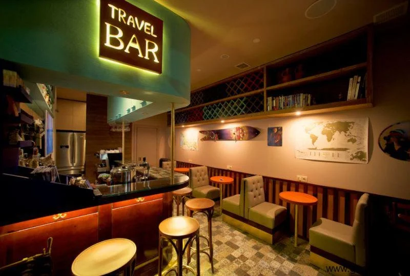 Interior of a travel-themed bar with a illuminated sign reading 'Travel Bar', cozy seating with green and beige chairs, wooden tables, and shelves filled with books and decor on the wall.