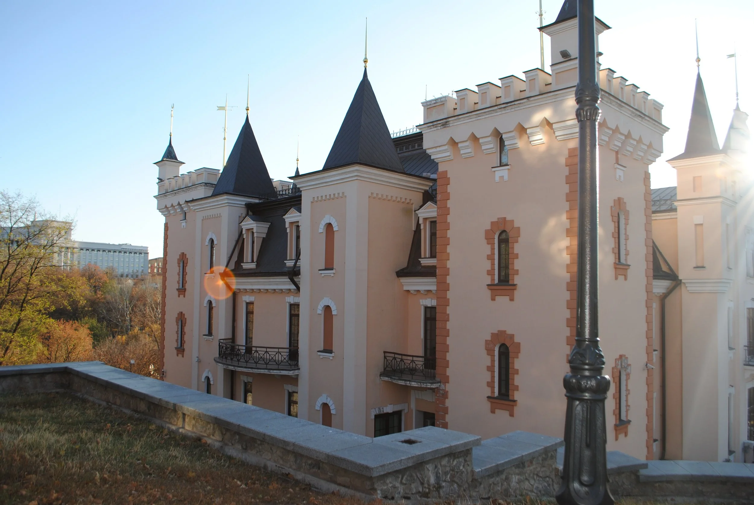 A historic castle-like building with turrets, beige walls, and decorative white and brick accents, set against a clear blue sky in a city park area.