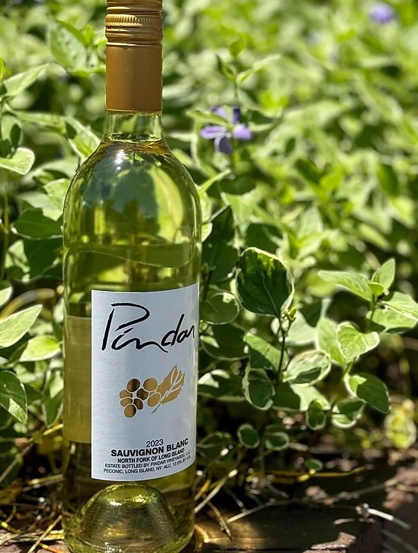 A clear glass bottle of white wine with a gold cap placed among green foliage and purple flowers.
