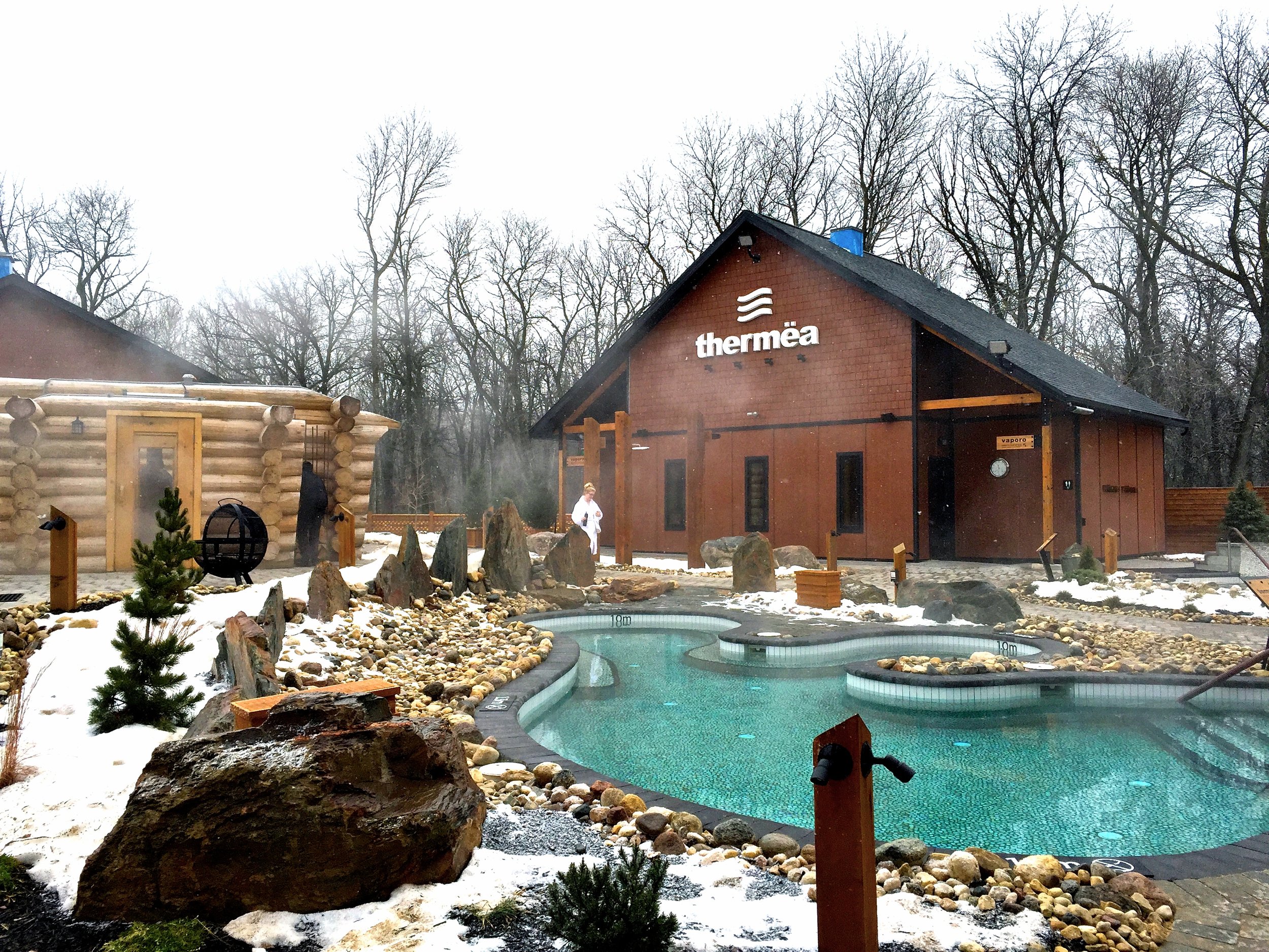 Snow-covered landscape with mountains, trees, and a modern outdoor hot spring pool complex with a cone-shaped glass structure.