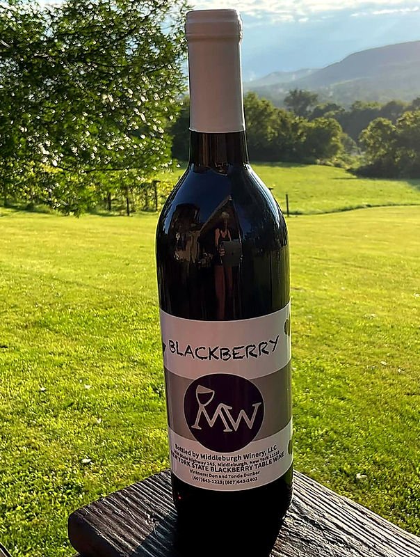 A bottle of homemade blackberry wine on a wooden surface outdoors with grass, trees, and mountains in the background.
