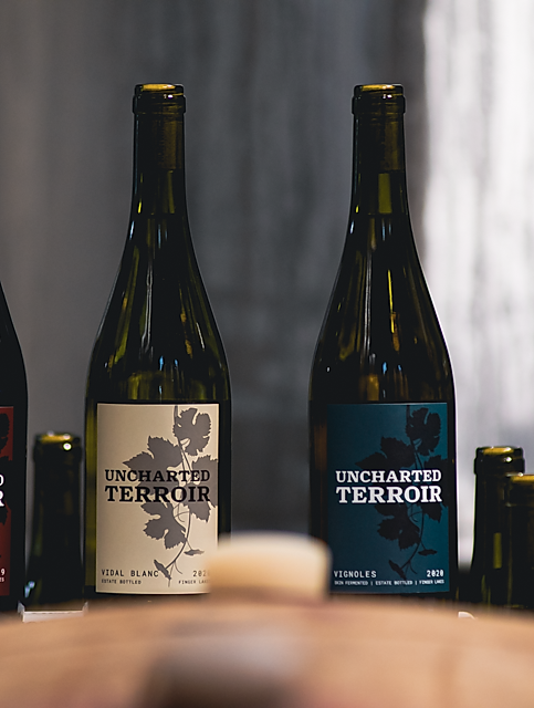 Two bottles of Uncharted Terroir wine, one Vidal Blanc and one Vignoles, with black labels and leaf illustrations, placed on a table with blurred bottles in the background.
