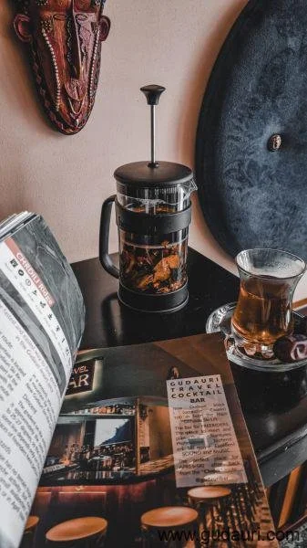 French press with a glass container filled with coffee or tea, placed on a black table next to a glass cup with tea, a magazine, and a small decorative cup. A wall with a mask and a large circular object are in the background.
