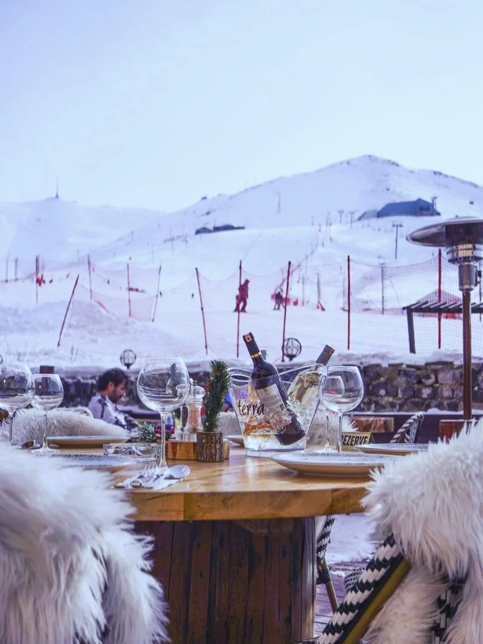 Indoor dining table set with wine bottles, glasses, and festive decorations, overlooking a snowy mountain landscape with skiers and ski lifts.