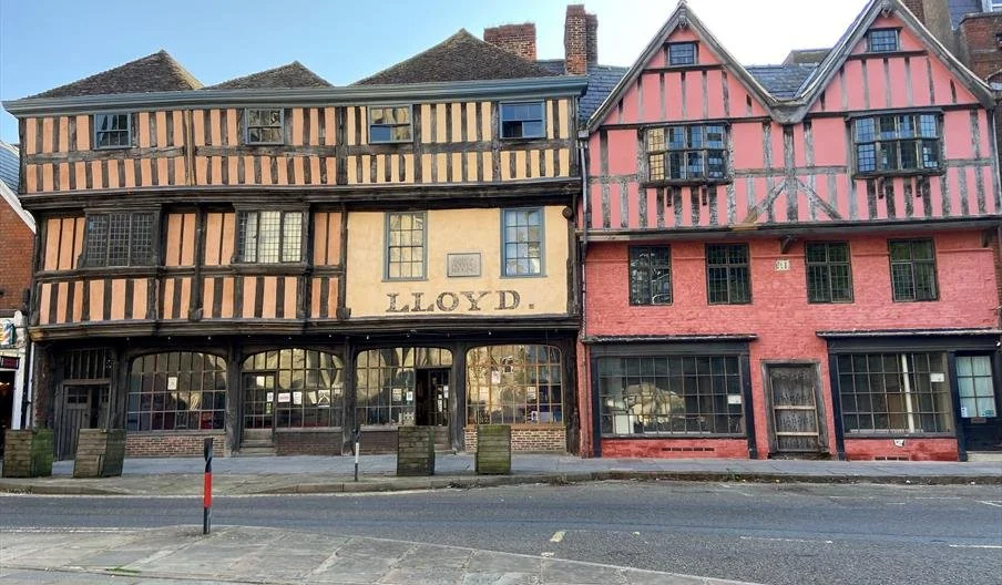 Two colorful historic buildings with Tudor-style timber framing; one has a beige and black pattern with the name 'LLOYD' on it, and the other is painted red with black timber accents.