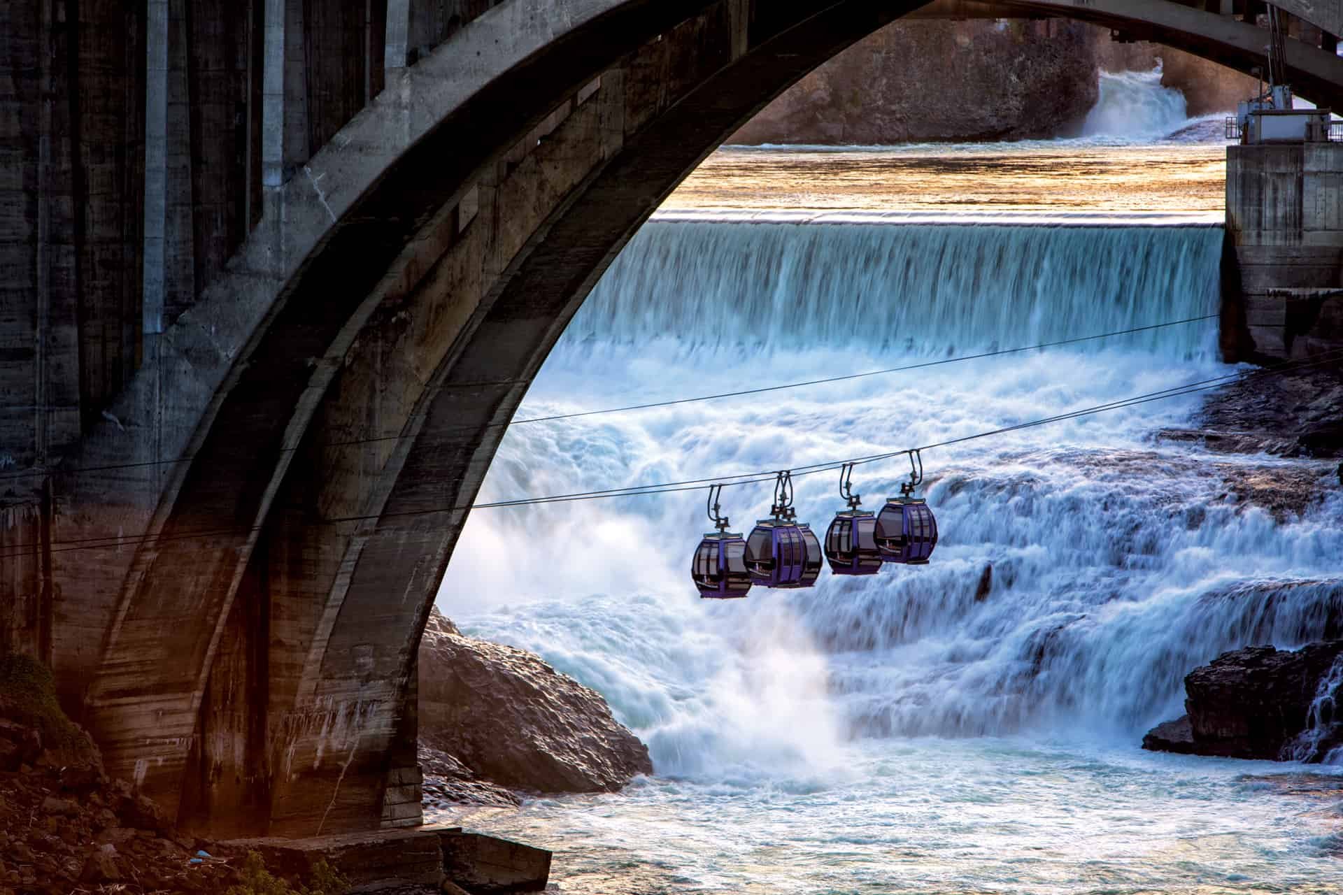 A dam with water flowing over it, and three gondolas on a cable above rushing water.
