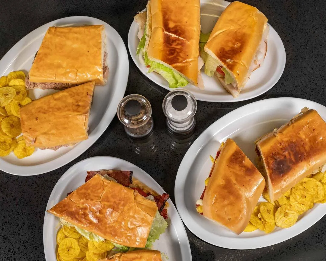 Four plates of sandwiches with sliced bread, lettuce, tomatoes, cheese, and meats on a black table, accompanied by yellow potato chips and salt and pepper shakers.