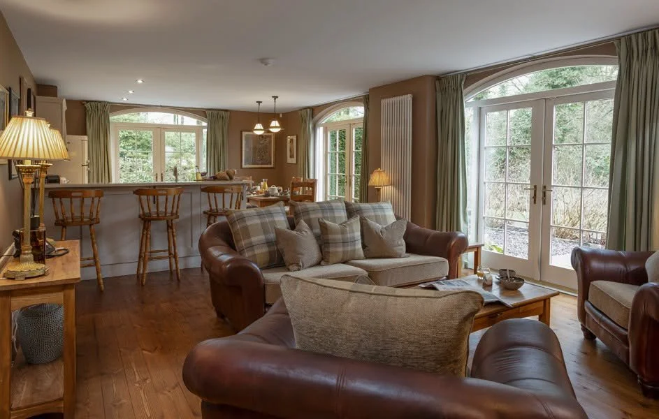 A cozy living room with leather and fabric sofas, a wooden coffee table, and large windows with green curtains, overlooking a garden.