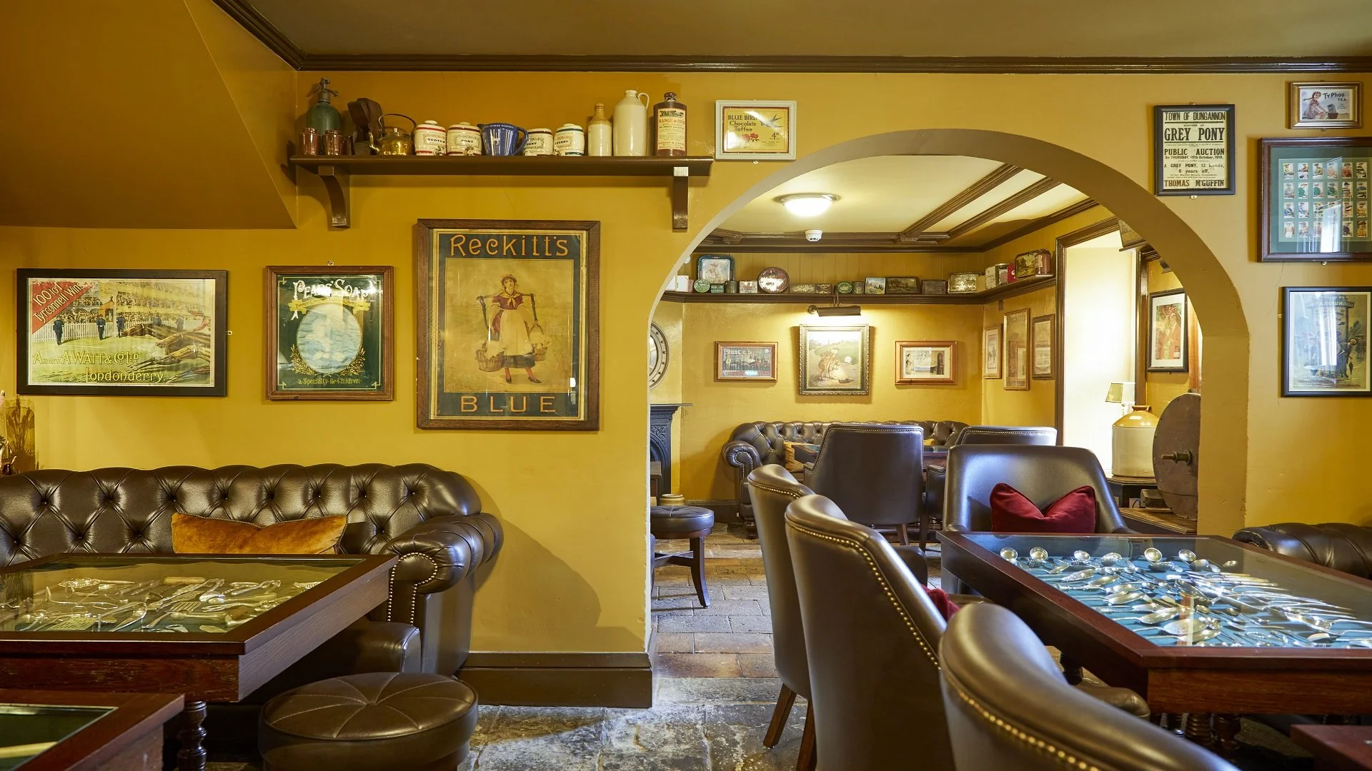 Interior of a cozy pub or restaurant with yellow walls, leather armchairs, framed vintage posters, and a glass-top table with silverware. There is an arched doorway leading to more seating area.
