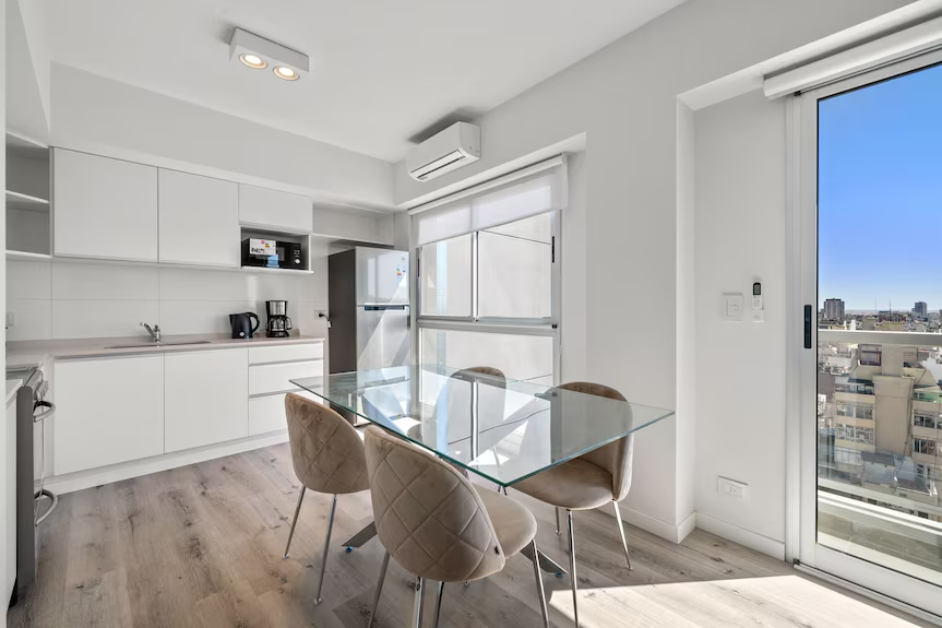 Modern kitchen and dining area with a glass table and four beige quilted chairs, white cabinets, microwave, coffee maker, refrigerator, large windows with city view, and a sliding glass door leading to a balcony.