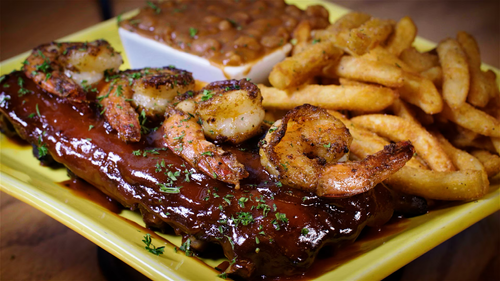 Barbecue ribs topped with barbecue sauce, grilled shrimp garnished with herbs, seasoned French fries, and baked beans on a yellow plate.