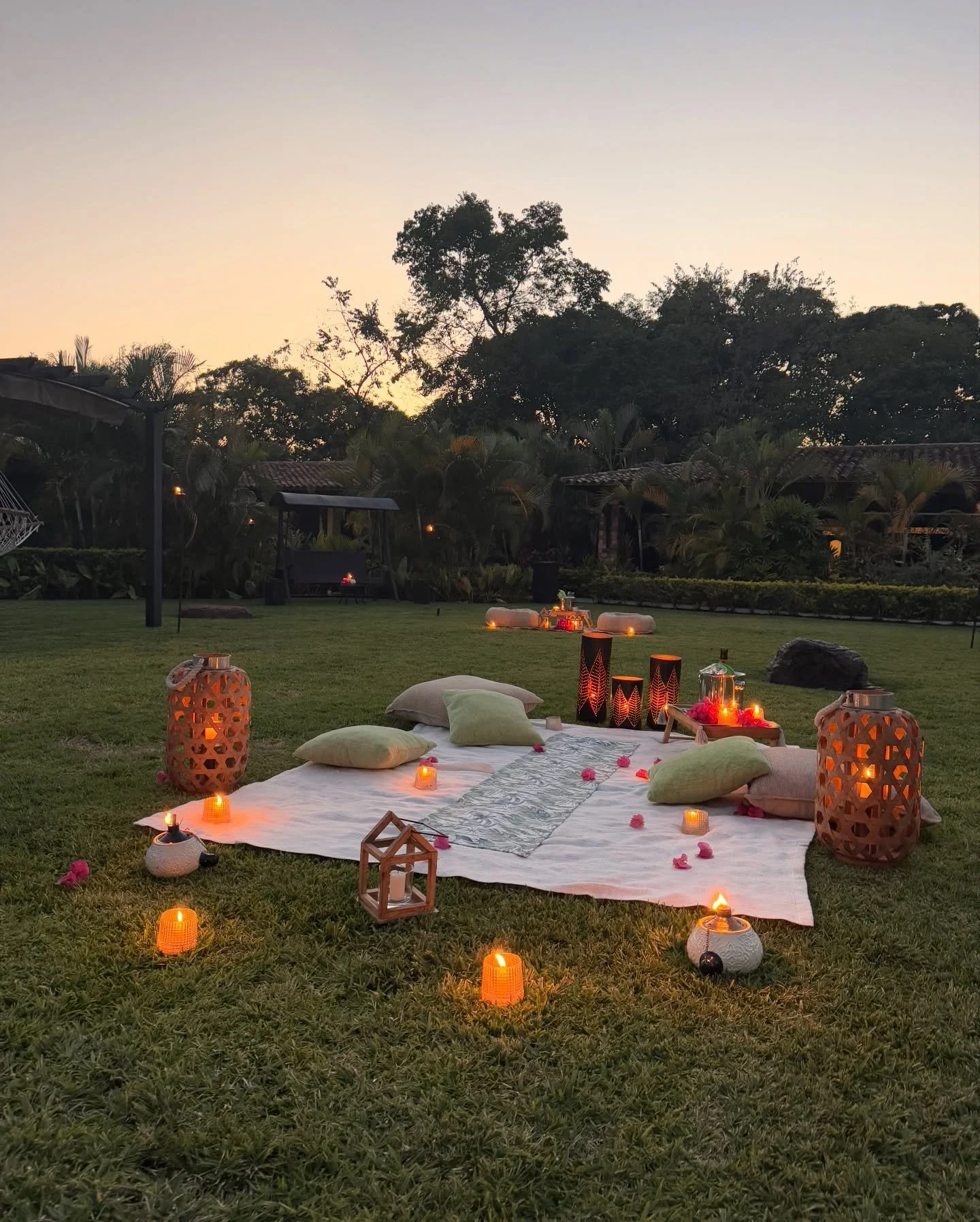 Outdoor romantic setup with a white blanket on grass, surrounded by candles, lanterns, pillows, and decorative items at sunset, with trees and rooftops in the background.