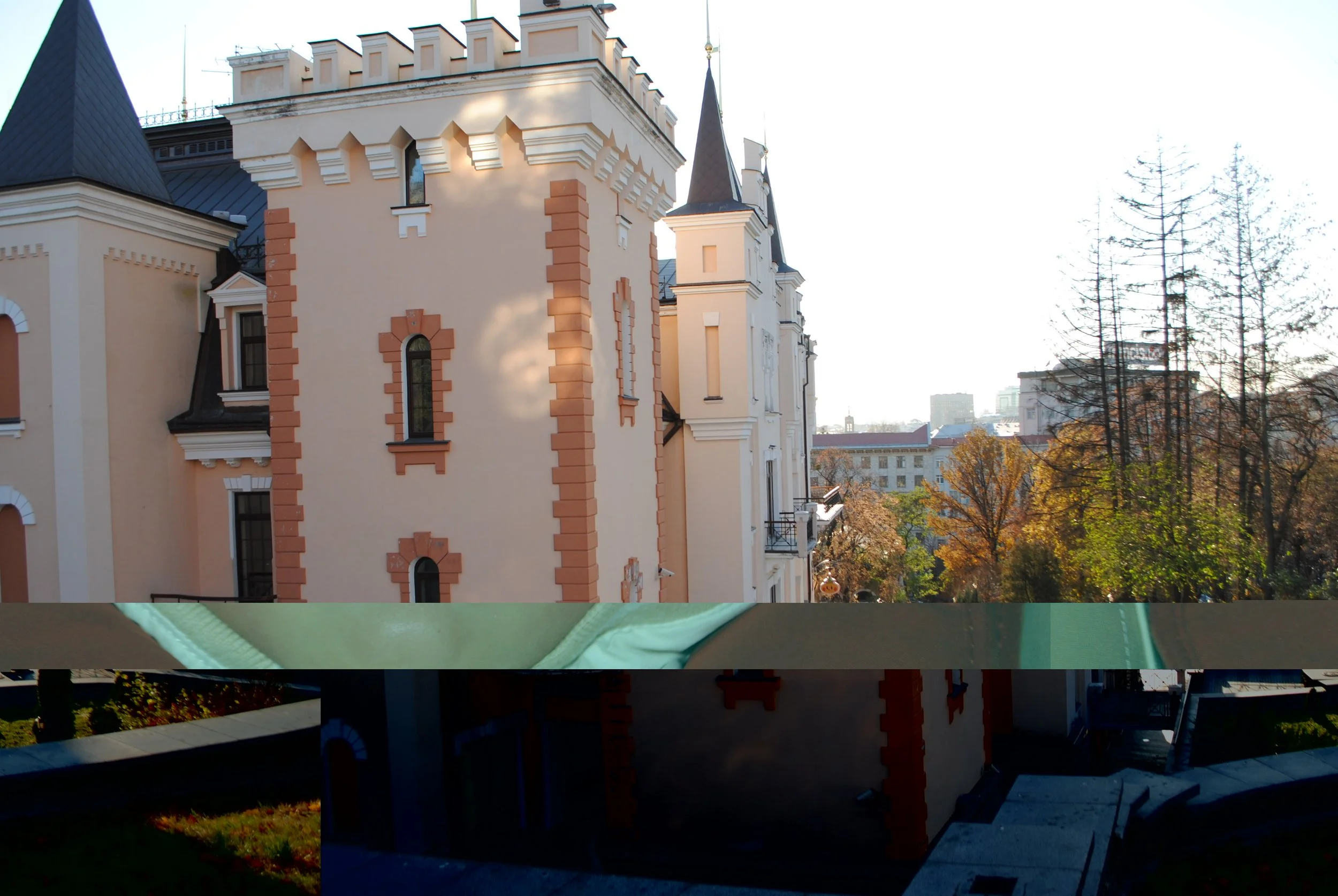 A castle-like building with beige walls and decorative brick accents, featuring narrow arched windows and turrets, set against a backdrop of trees with yellow autumn leaves and a distant cityscape.