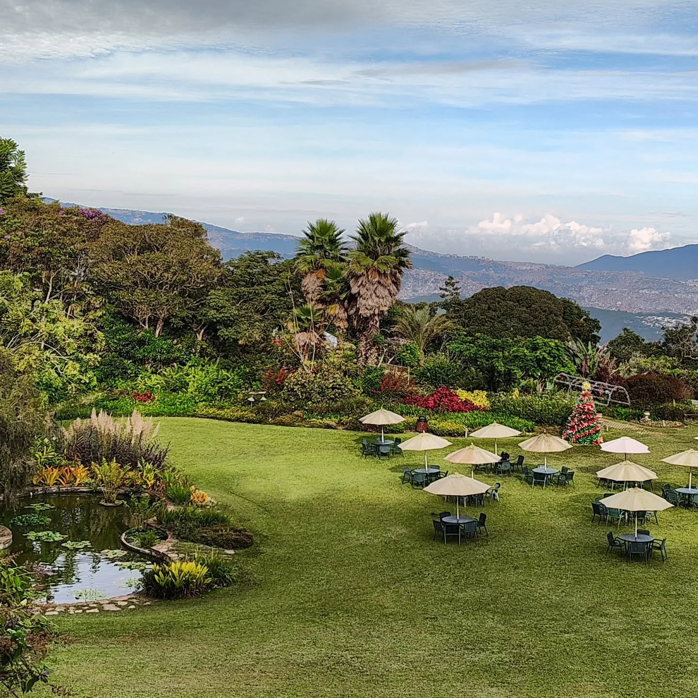 A lush garden with a pond, surrounded by trees and colorful flowers, with restaurant tables and umbrellas on a well-maintained lawn, mountains and a partly cloudy sky in the background.