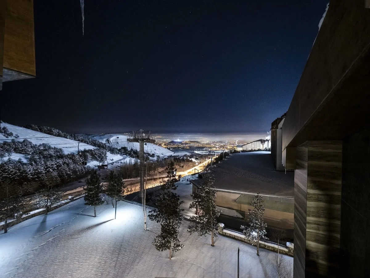 Nighttime view of snow-covered slopes, trees, and a distant cityscape with lights under a clear starry sky, seen from a balcony or raised platform with dark wooden exterior.