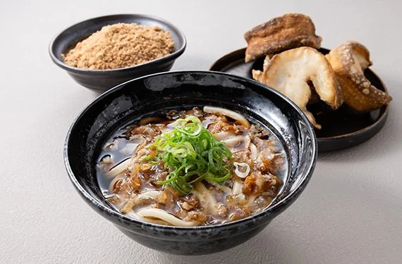 A bowl of Japanese beef and onion sukiyaki with green onions on top, a dish of ground sesame seeds, and a plate of fried tofu slices on a light gray surface.