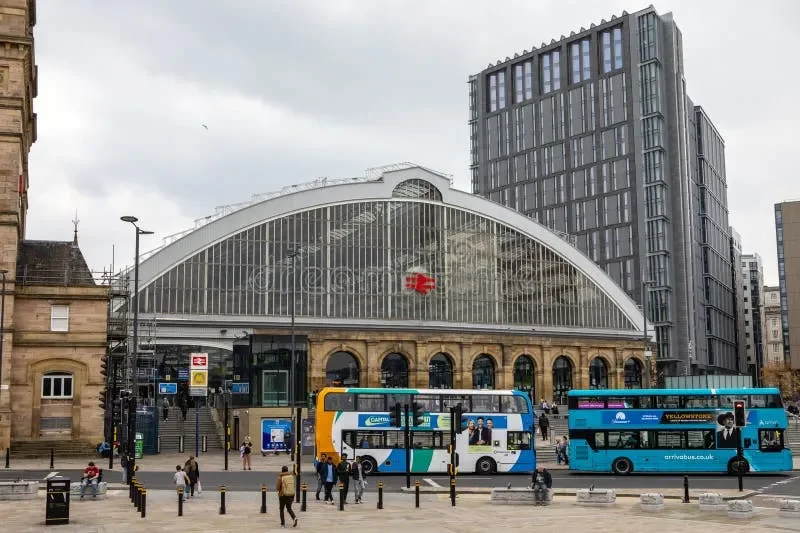 Exterior view of a large train station with historic architecture, glass roof, and modern high-rise buildings nearby, featuring buses and pedestrians outside.