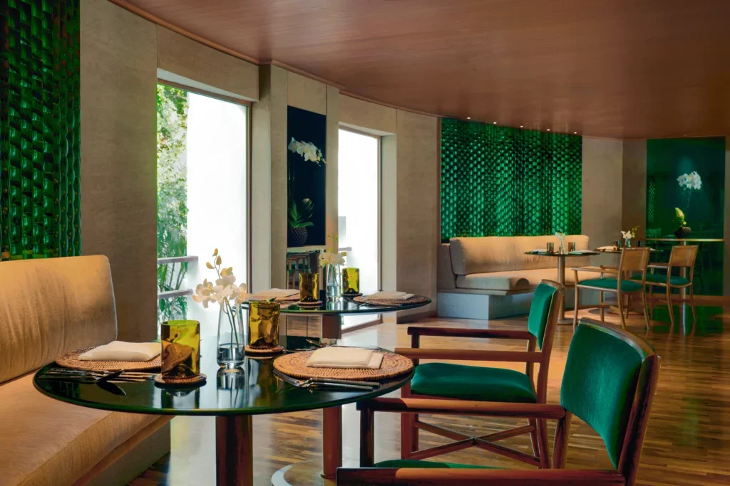 Interior of a modern restaurant with tables set with napkins, glasses, and cutlery, accented by green and beige decor, with large windows and green textured wall panels.
