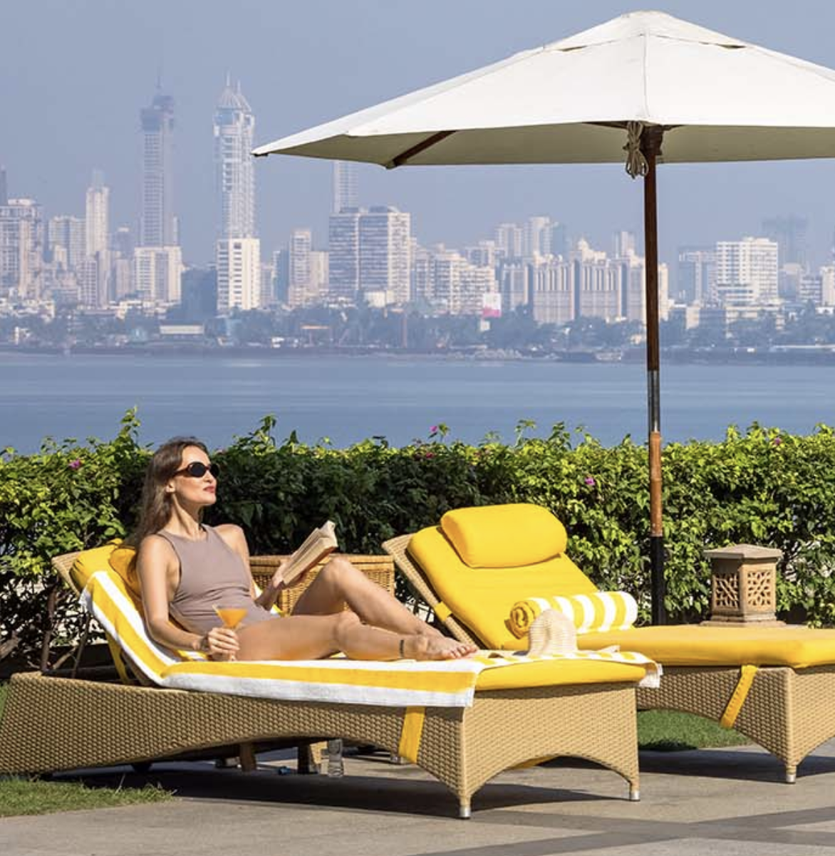 Woman relaxing on a yellow lounge chair with a drink and reading a book under a large white umbrella, with city skyline and water in the background.