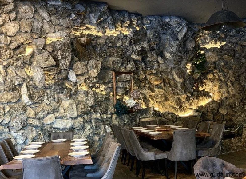 Interior of a restaurant with a rustic stone wall, dining tables set with plates, and upholstered chairs.