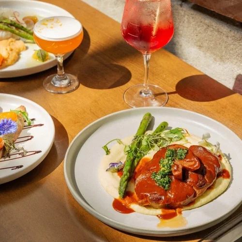 A plate of food with mashed potatoes, asparagus, a piece of meat with sauce, garnished with herbs. In the background, there are two glasses of colorful beverages, one orange and one red. Part of another plate with a salad or appetizer is visible on the left.