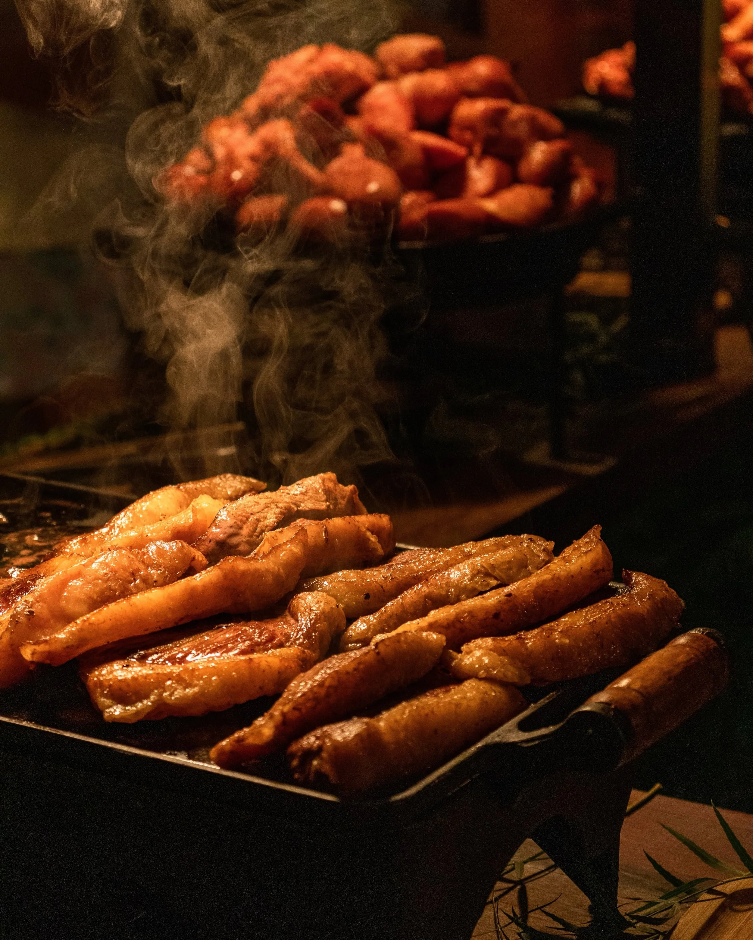 Grilled chicken strips on a black grill with steam rising, a tray of cooked chicken in the background, and some grilling tools visible.