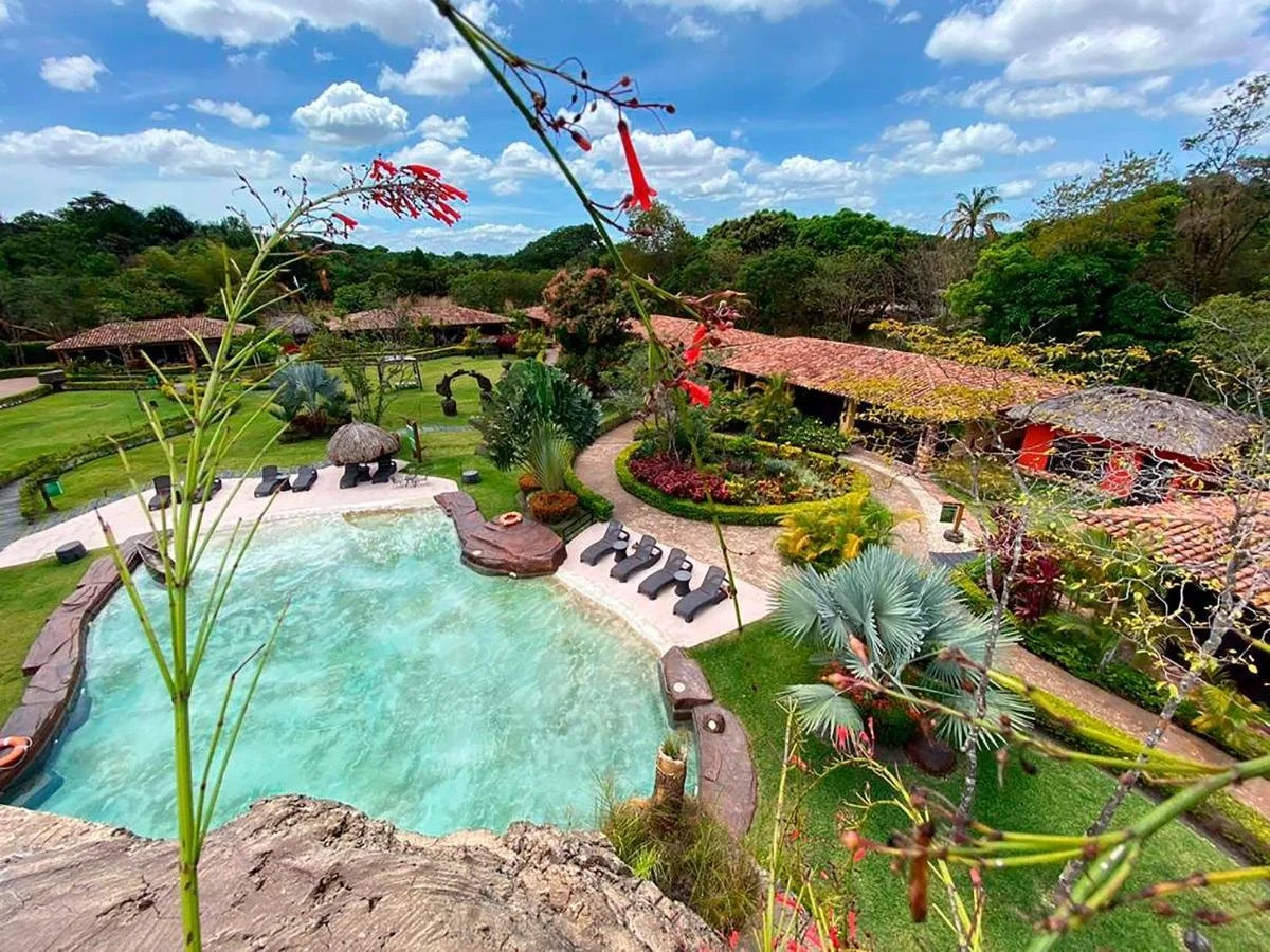 View of a tropical resort with a large swimming pool, lounge chairs, lush greenery, flowering plants, and rustic buildings with tiled roofs under a partly cloudy sky.