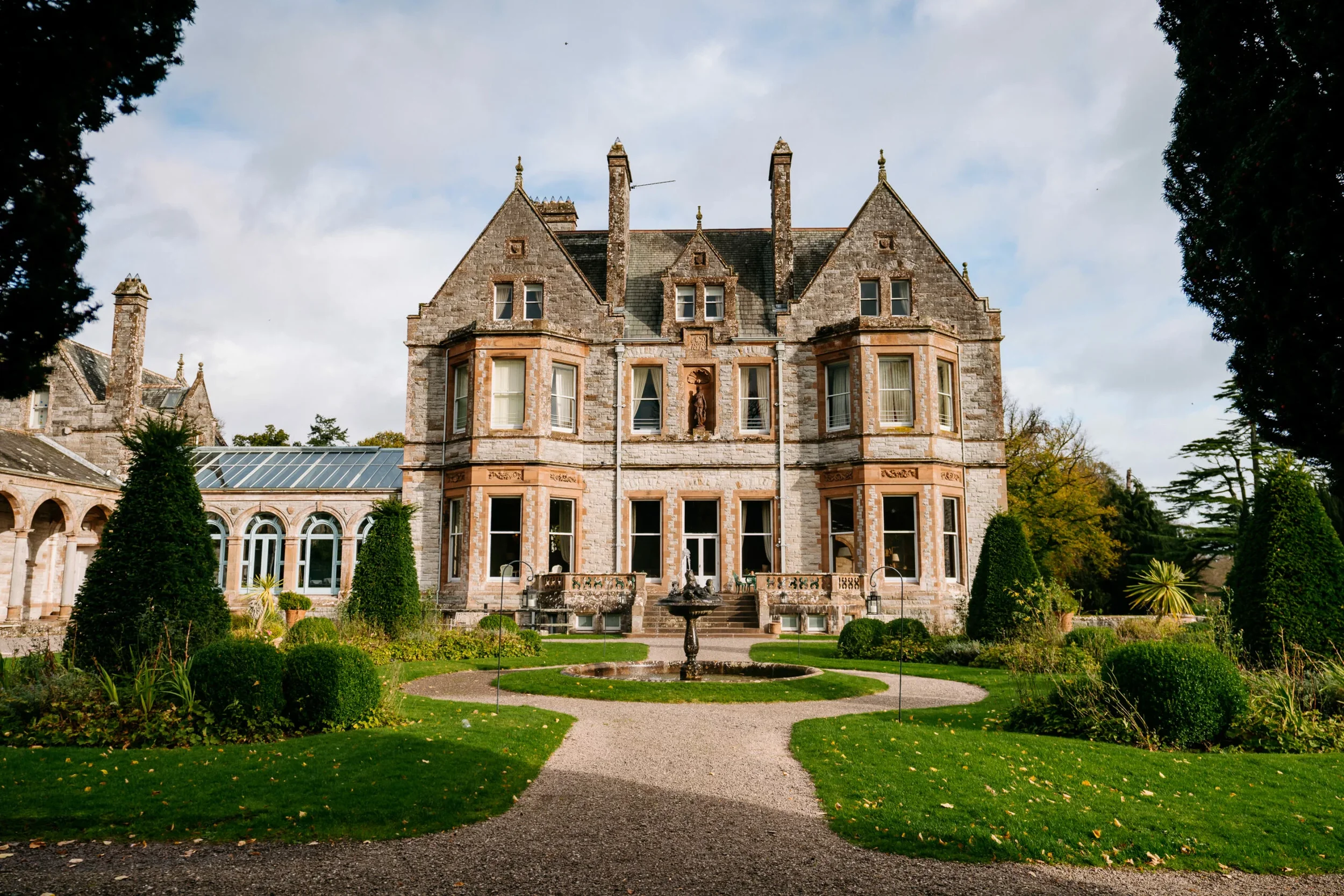 A large historic stone mansion with multiple gables, chimneys, and arched windows, surrounded by a landscaped garden with a fountain and a gravel pathway leading to the front entrance.